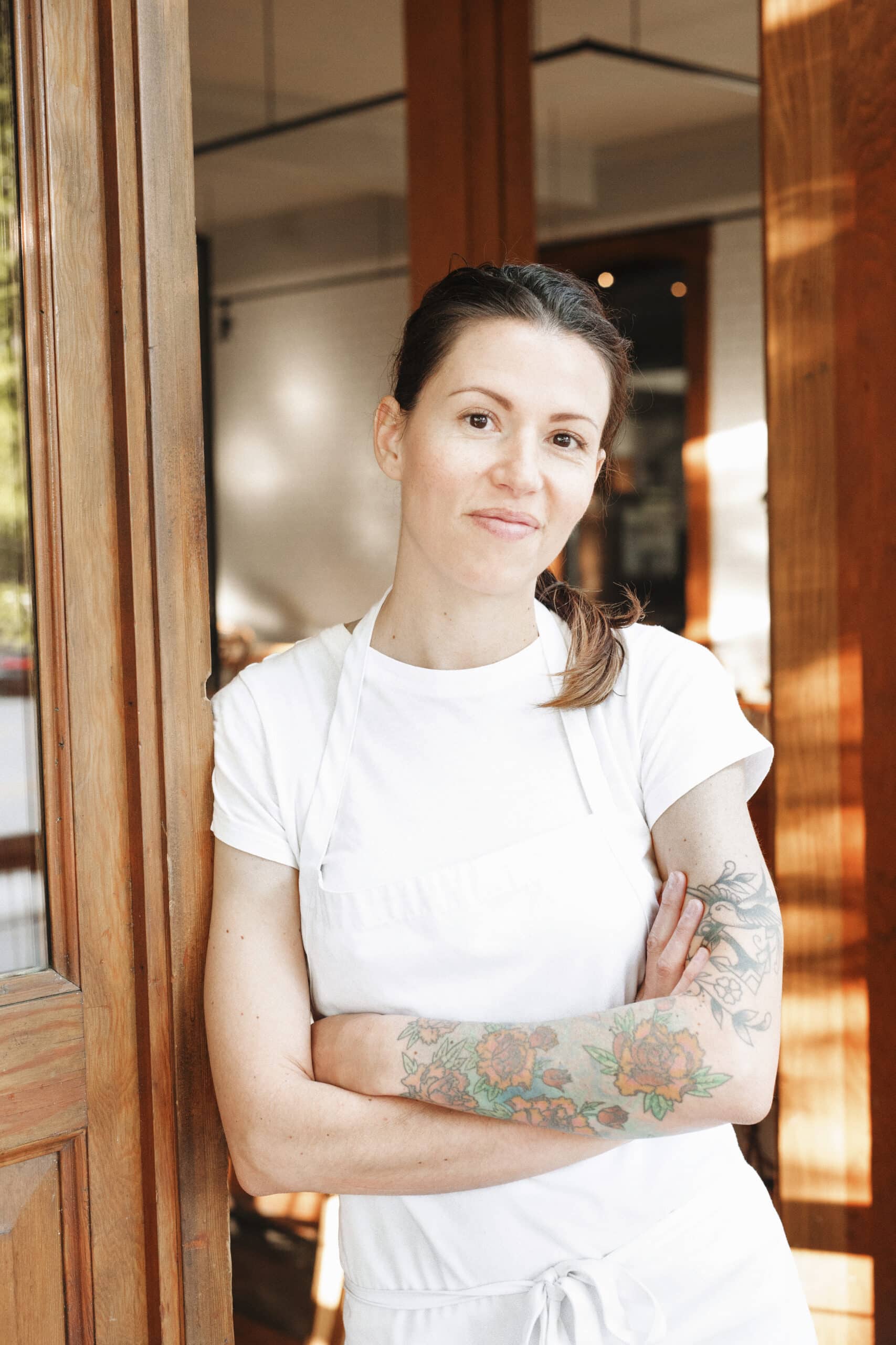 Portrait of a woman wearing a white apron standing with her arms crossed in a doorway, tattoos on her left arm. ,Lifestyle,USA